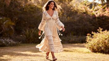 Mulher brasileira sorridente usando vestido de crochê longo e delicado em tom claro, caminhando ao ar livre sob luz natural.