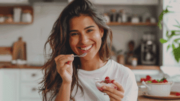 Mulher sorridente com pele iluminada saboreando iogurte com frutas vermelhas em uma cozinha clara, representando o cuidado com a saúde e a beleza da pele por meio da alimentação equilibrada.