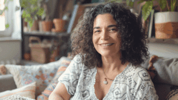 Mulher com sorriso confiante sentada em um sofá em uma sala iluminada e decorada com plantas, transmitindo serenidade e autoestima.