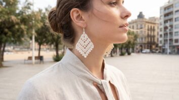 Mulher com brincos de crochê off-white em ambiente urbano veranil, design delicado com praia e prédios ao fundo