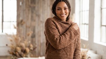 Mulher sorridente usando casaco de crochê artesanal em algodão marrom com gráficos geométricos, em ambiente outono/inverno com luz natural realçando elegância e sofisticação.