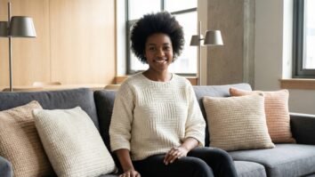 Mulher jovem sorridente sentada em sofá usando blusa de crochê moderna e criativa em algodão off-white com design minimalista, cercada por almofadas de crochê em cores coordenadas em ambiente urbano sofisticado.