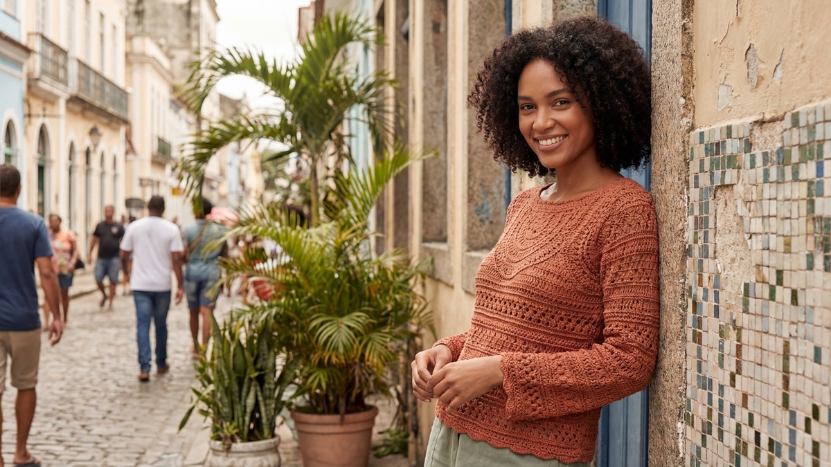 Mulher sorridente usando blusa de crochê artesanal em tom terracota, em ambiente urbano de verão com luz natural difusa destacando a textura e caimento do crochê.