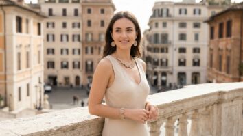 Mulher jovem elegante apoiada em balaústre de pedra clara, usando vestido sem mangas bege/creme claro com decote em V, acessórios artesanais de crochê em tons bege/creme incluindo brincos circulares detalhados, colar gargantilha com padrão intrincado em formato de vieiras ou flores, e pulseira circular.