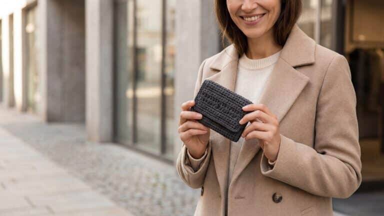Mulher sorridente segurando carteira de crochê artesanal cinza grafite com textura moderna, composição editorial minimalista em cenário urbano com iluminação natural.