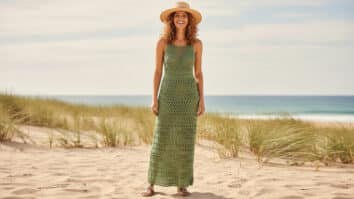 Mulher de cabelo cacheado sorrindo com vestido de crochê artesanal em algodão verde claro, modelagem elegante e leve, acompanhada de chapéu minimalista e sandália neutra, em cenário veranil de praia com luz natural difusa.