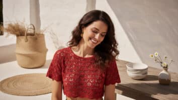 Mulher morena sorrindo usando cropped de crochê vermelho com padrão estiloso em ambiente com luz natural e elementos decorativos sutis