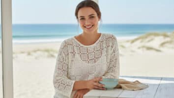 Mulher jovem sorrindo usando blusa de crochê leve em fio de algodão off-white com padrão filet geométrico arejado, em ambiente iluminado com elementos discretos de linho e cerâmica clara, destacando o design vazado e o caimento fresco da peça em atmosfera veraneia.