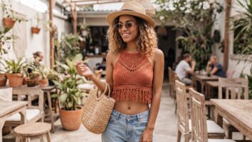 Mulher brasileira sorridente em top crop de crochê artesanal terracota com barriga à mostra, óculos minimalistas, chapéu de palha e jeans dobrado em estilo contemporâneo.