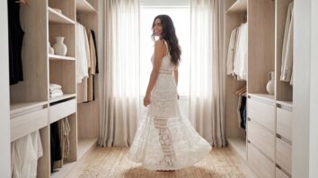 Mulher sorridente em closet minimalista usando vestido artesanal de crochê em algodão branco, estilo romântico e sofisticado, com luz natural difusa revelando o caimento, movimento e trama rendada delicada da peça.