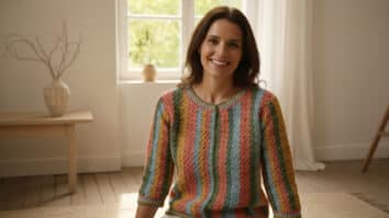 Mulher sorridente usando blusa de crochê colorida em ambiente naturalista com iluminação suave, decoração minimalista com vaso de cerâmica e elementos secos