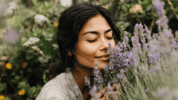 Imagem de uma mulher com olhos fechados cheirando um ramo de lavanda em um campo florido