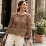 Mulher sorrindo em ambiente urbano usando blusa de crochê marrom elegante com pontos abertos e textura sofisticada.