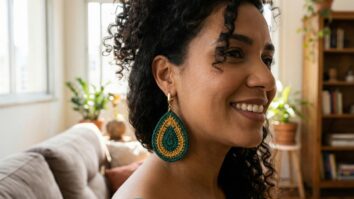 Mulher brasileira sorrindo em ambiente urbano iluminado, usando brincos de crochê em formato de gota nas cores verde esmeralda e mostarda.