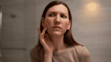 Mulher analisando o rosto no espelho, onde há vermelhidão nas bochechas, um possível sinal de infamação