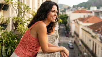 Mulher brasileira sorrindo em ambiente urbano de verão, vestindo top de crochê artesanal coral com alças delicadas.