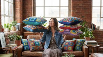 Ambiente urbano casual com mulher sorrindo mostrando almofadas de crochê artesanal finalizadas em squares geométricos coloridos, confeccionadas em fio de algodão, com pontos fechados e acabamento firme