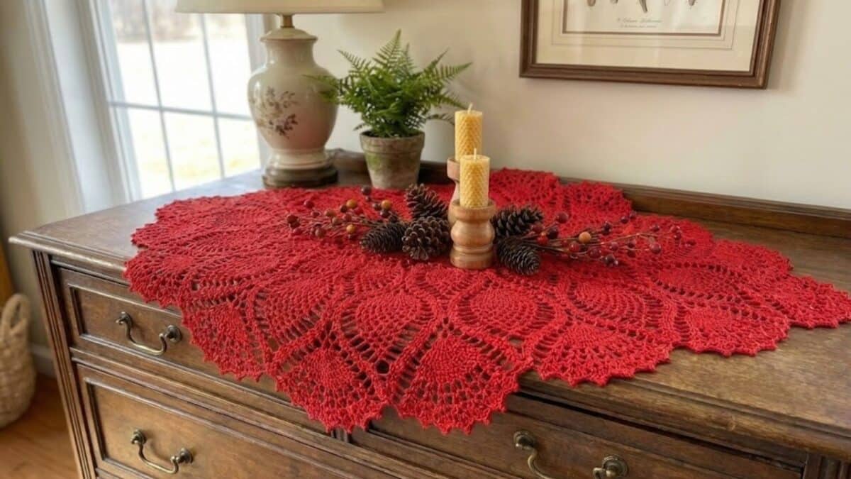Um centro de mesa de crochê vermelho com um padrão de leque está sobre uma cômoda de madeira antiga. No centro do crochê, há um arranjo com duas velas em castiçais, pinhas e ramos de frutas vermelhas. Atrás dele, sobre a cômoda, estão um abajur, um pequeno vaso com uma samambaia e um quadro emoldurado em ambiente rústico e aconchegante.