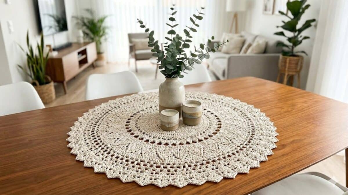 Centro de mesa em crochê redondo na cor crua posicionado sobre uma mesa de jantar de madeira em uma sala moderna e iluminada, decorado com um vaso de cerâmica contendo ramos de eucalipto e duas velas pequenas.