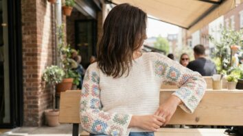 Ambiente urbano leve e contemporâneo apresentando mulher sorrindo usando blusa de crochê artesanal finalizada com granny squares, modelo moderno e feminino, corpo da blusa liso em ponto fechado enquanto as mangas são compostas por granny squares coloridos unidos com precisão
