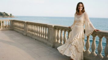 Cena editorial de moda sofisticada em ambiente externo elegante com mulher sorrindo usando vestido de crochê artesanal em estilo boho-romântico, apresentando babados delicados na saia e mangas ciganinha amplas