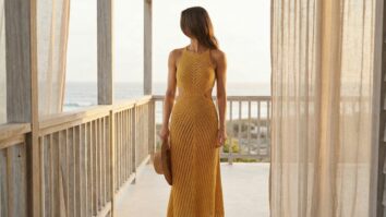 Ambiente de verão sofisticado de varanda elegante ou cenário costeiro minimalista, apresentando mulher sorrindo usando vestido de crochê artesanal finalizado modelo longo e fluido com recortes laterais (cut-out) na cintura, em tom amarelo charmoso