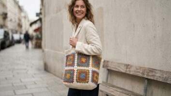 Mulher sorrindo em estilo street style urbano usando bolsa de crochê artesanal com squares geométricos multicoloridos sobre base em tons naturais de algodão cru, criada em fio de algodão, com destaque para a peça como statement piece bohemian-chic contemporâneo