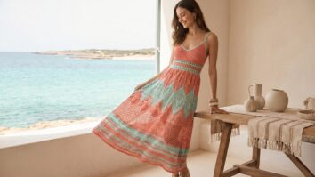 Terraço à beira-mar no verão com mulher sorrindo usando vestido longo de crochê em tons vibrantes de verão (coral e turquesa suave).