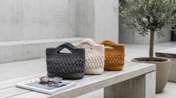 Ambiente urbano contemporaneo com bolsas artesanais de croche em cores modernas e sofisticadas dispostas sobre banco de madeira clara ao lado de oculos de sol elegantes e revista de moda fechada
