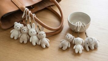 Chaveiros de croche artesanal em tons naturais off-white, bege e cru, pendurados como bag charms em bolsa de couro neutra sobre superficie de madeira clara, em cenario boho chic com iluminacao natural difusa