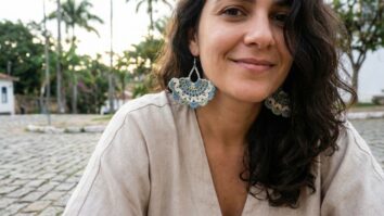 Mulher sorrindo usando brincos artesanais de croche em tons coloridos com trama rendada em ponto leque, ambiente externo com luz natural
