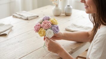 Mesa de madeira clara com mulher segurando flores de croche artesanal em tons rosa, amarelo, branco e lavanda
