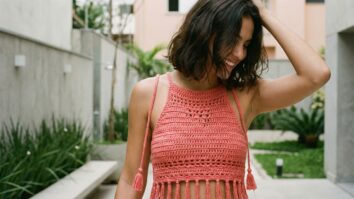 Mulher sorrindo usando blusa de croche artesanal vermelho claro em ambiente urbano brasileiro, estilo moderno e elegante com luz natural