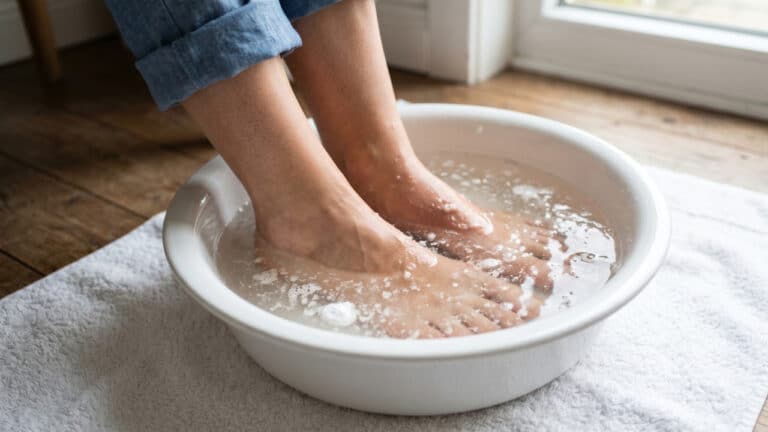 Pés submersos em uma bacia branca com água morna, cristais de sal grosso e bicarbonato de sódio visíveis no fundo, posicionada sobre uma toalha branca em um piso de madeira.