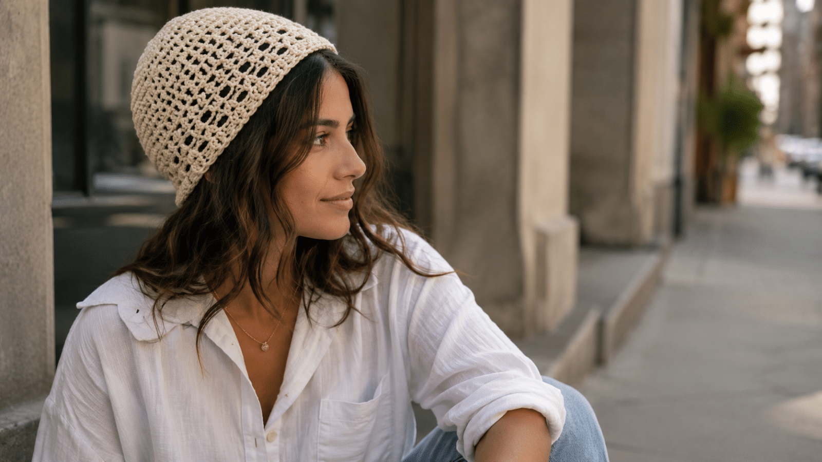 Mulher usando touca de crochê moderna em tom neutro, com look casual de camisa branca e jeans em ambiente urbano.