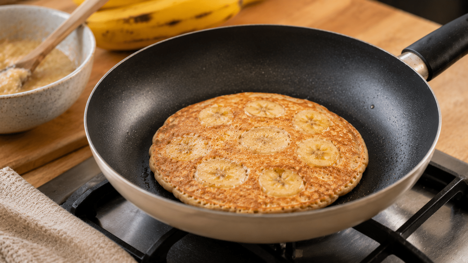 Panqueca de banana sendo preparada na frigideira, com rodelas da fruta incorporadas à massa de forma natural e dourada.