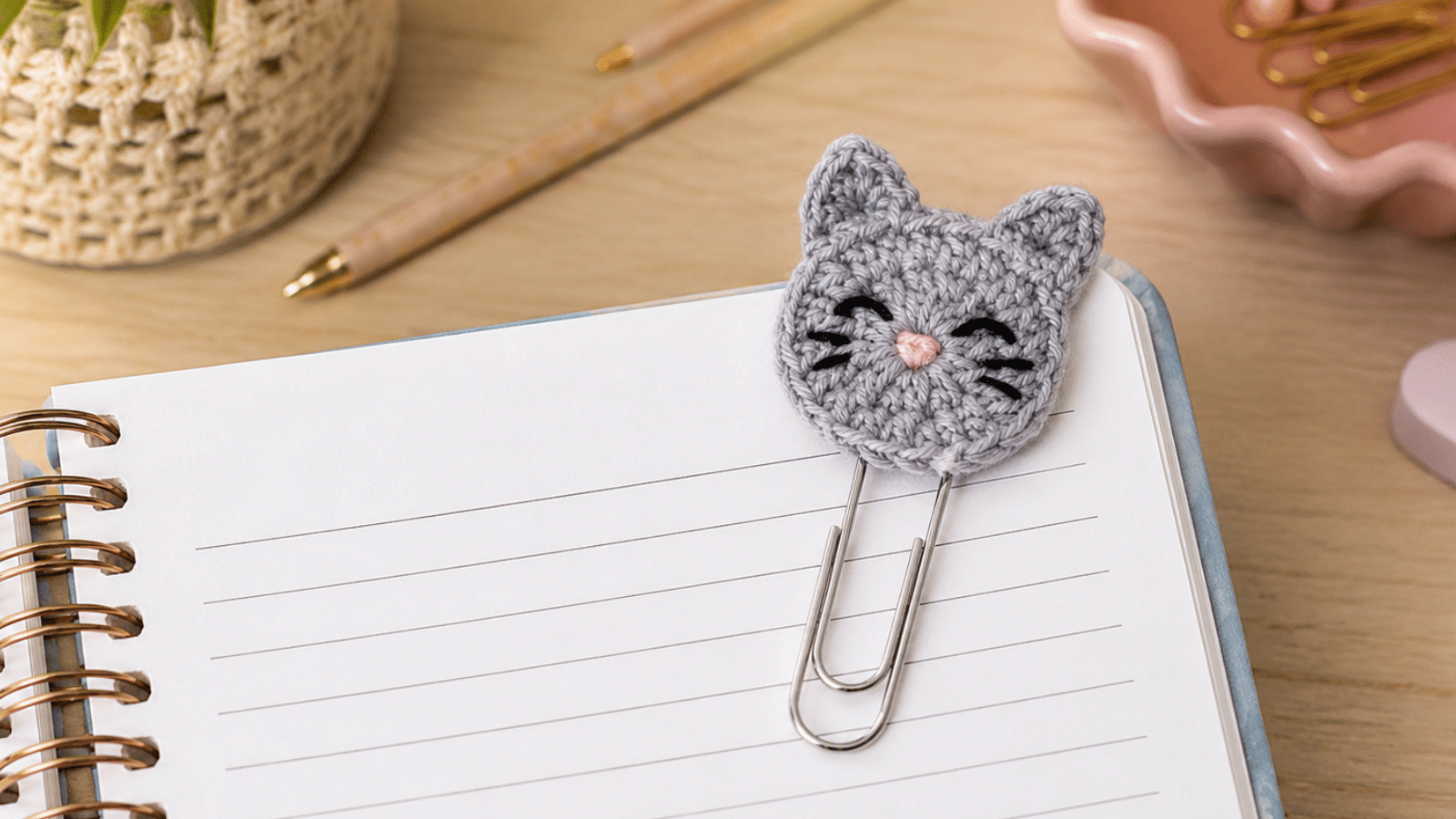 Clipe de papel com gatinho em crochê sendo usado como marcador em caderno, destacando a delicadeza artesanal e o charme da papelaria criativa.