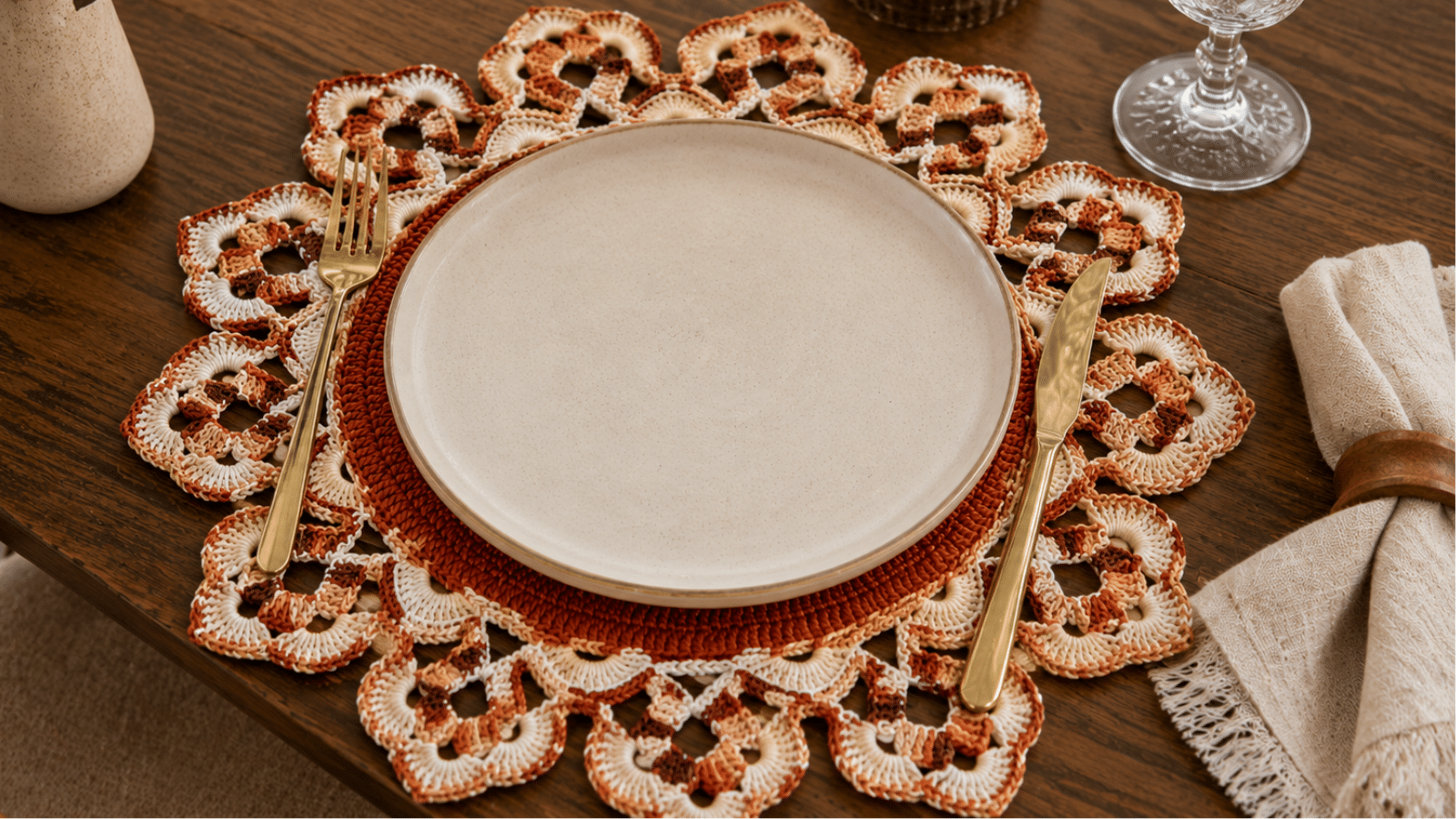 Sousplat de crochê rendado em tons terrosos decorando mesa posta com prato neutro, talheres dourados e composição acolhedora.