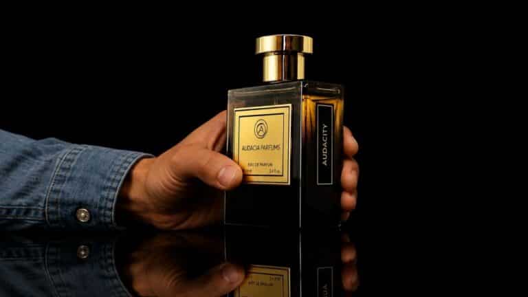 Perfume Audacity da Azza Parfums sendo segurado por uma mão masculina sobre superfície espelhada, fragrância que viralizou entre os fãs de perfumaria em 2026.