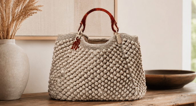 Bolsa de crochê bege com textura em ponto pipoca, alças marrons arredondadas e acabamento artesanal, exposta sobre uma superfície de madeira em ambiente neutro e elegante.