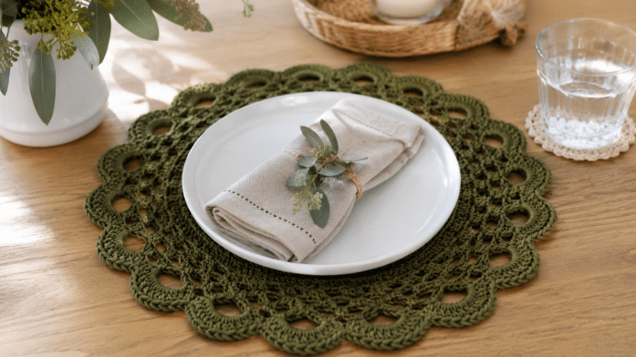 Sousplat de crochê verde-oliva com borda rendada e acabamento arredondado, usado em uma mesa posta com prato branco, guardanapo de tecido e decoração natural.