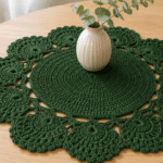 Toalha de mesa de crochê verde com centro circular fechado e bordas florais vazadas, decorada com vaso pequeno sobre mesa de madeira clara em ambiente iluminado.