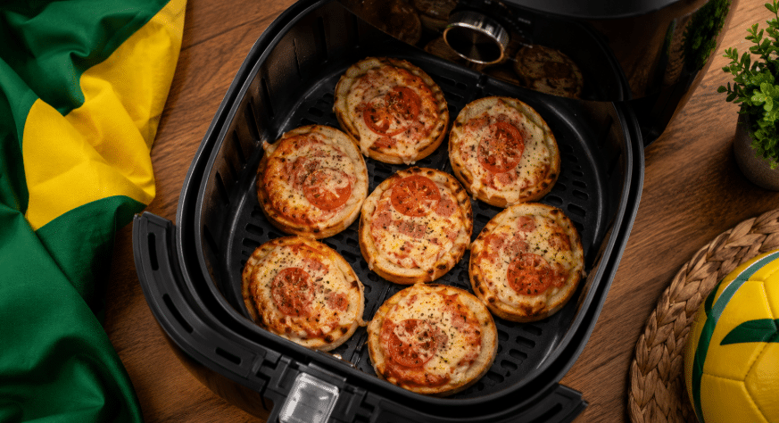 Mini pizzas caseiras dentro de uma Air Fryer aberta, vistas de cima, com queijo derretido, tomate, orégano e detalhe em verde e amarelo ao lado remetendo ao Brasil.
