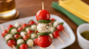 Espetinho caprese em close, com tomate-cereja, muçarela e folhas de manjericão, segurado em destaque sem mostrar a mão, com outros espetinhos desfocados ao fundo.
