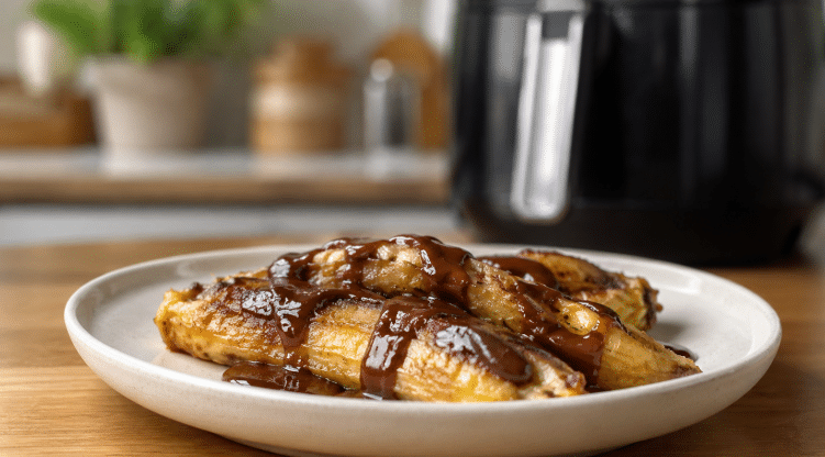 Bananas assadas na Air Fryer com cobertura de chocolate derretido, servidas em prato claro sobre mesa de madeira, com a fritadeira ao fundo desfocada.