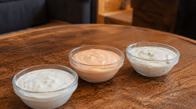 Três potinhos de molhos caseiros sobre uma mesa rústica de madeira, incluindo um molho branco de alho, um molho rosé e um molho claro com ervas.