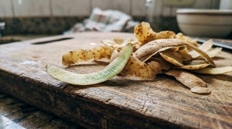 Fitas de casca de batata crua sobre uma tábua de corte de madeira rústica. Em primeiro plano, uma fita inteira de casca chama a atenção por apresentar uma forte coloração verde na parte interna.