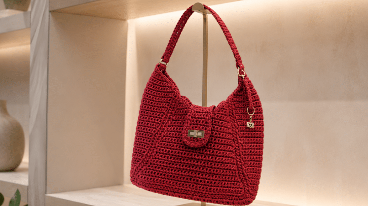 Bolsa de crochê em tom marsala profundo exposta em suporte metálico dourado, em ambiente de loja minimalista com iluminação suave e fundo neutro.