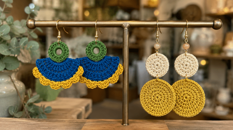 Dois pares de brincos de crochê expostos em um suporte de loja, sendo um modelo colorido em verde, azul e amarelo e outro modelo redondo em tons de bege e amarelo.