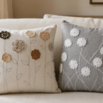 Duas almofadas decorativas com flores de crochê aplicadas, uma em tecido claro e outra cinza, apoiadas lado a lado sobre um sofá bege em ambiente aconchegante.
